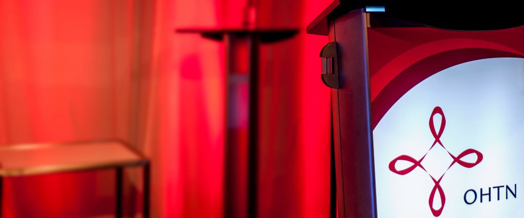 A podium with the OHTN logo on it stands before a curtain, chair, and table in dramatic red lighting.