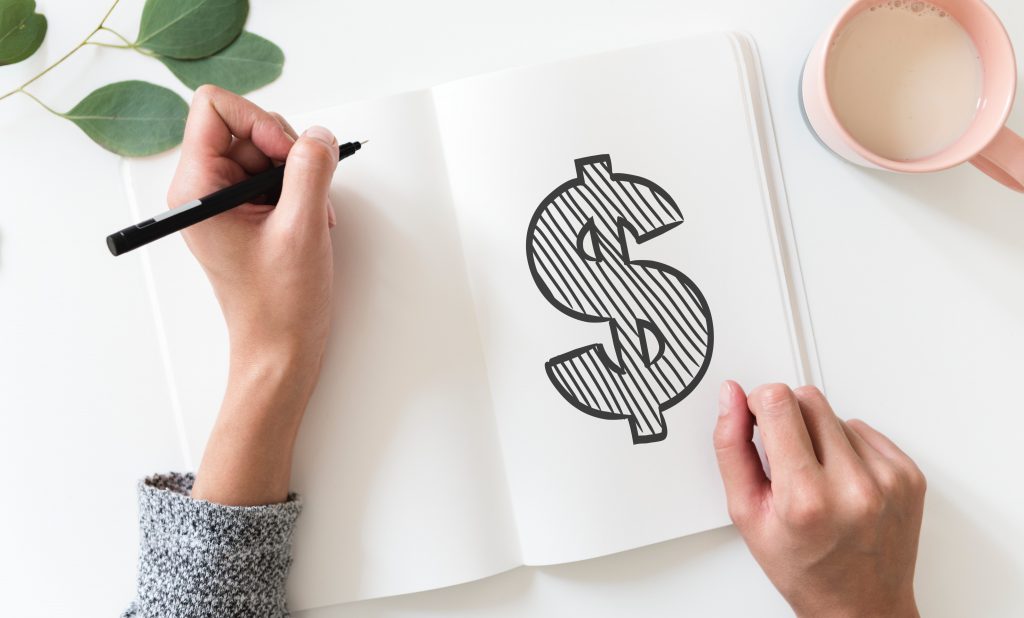 Bird's eye view of hands drawing a dollar sign in a sketchbook with a hot drink off to the side