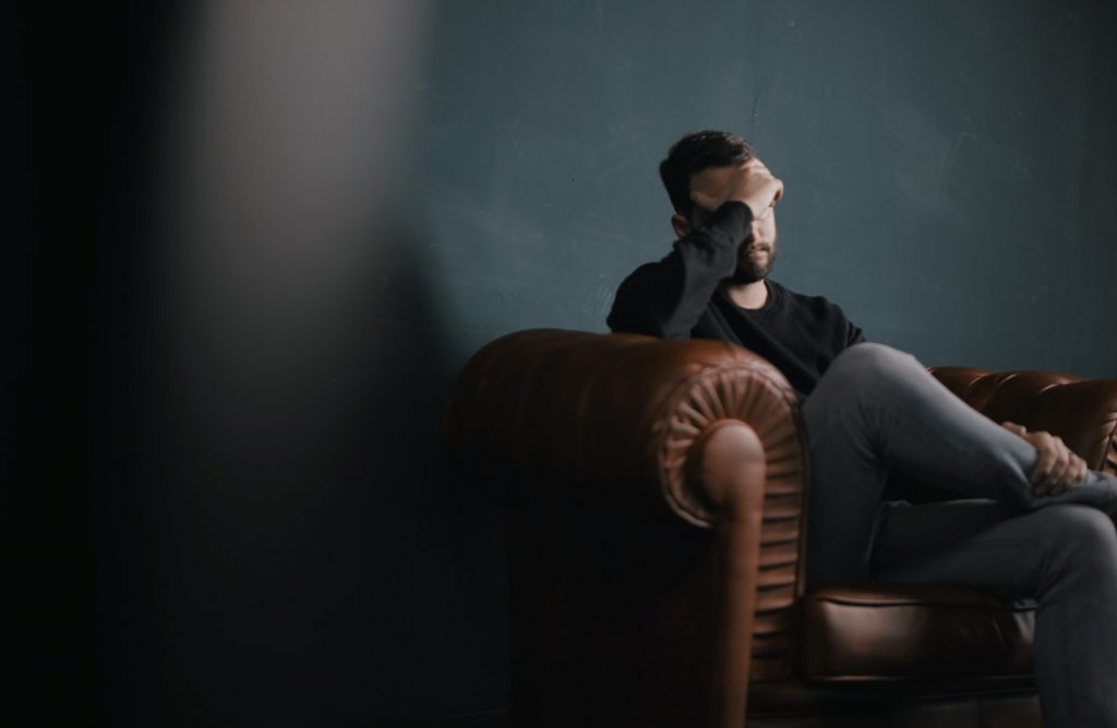 Man sitting on sofa against wall with his right hand covering his eyes and forehead