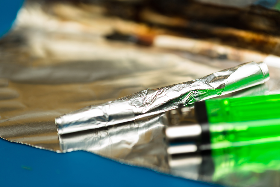 Authentic close up image of a rolled up foil tube used to smoke heroin, sitting on a used piece of foil next to a green disposable lighter, with melted heroin visible on the foil in the background, out of focus due to shallow depth of field.